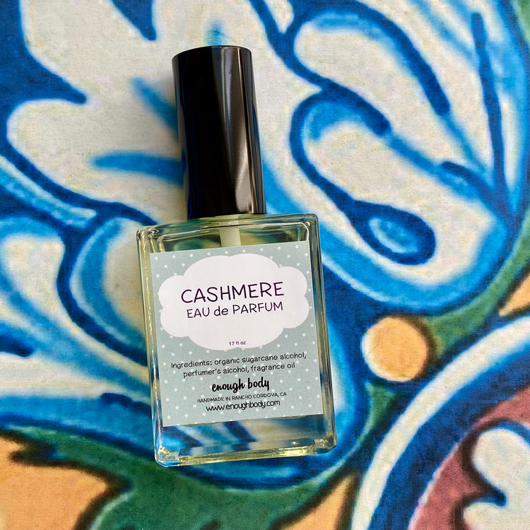 A glass bottle of Cashmere Eau de Parfum spray with a black spray mister top, labeled with the product name and a description as 'A feminine blend of blueberry, white freesia, wild grape, pomegranate, lily, magnolia, green vines, cashmere, and violet.'.