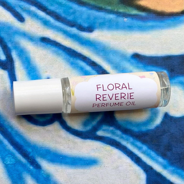 A glass bottle with a white cap labeled 'floral reverie Perfume Oil' set against a blue, floral patterned background.