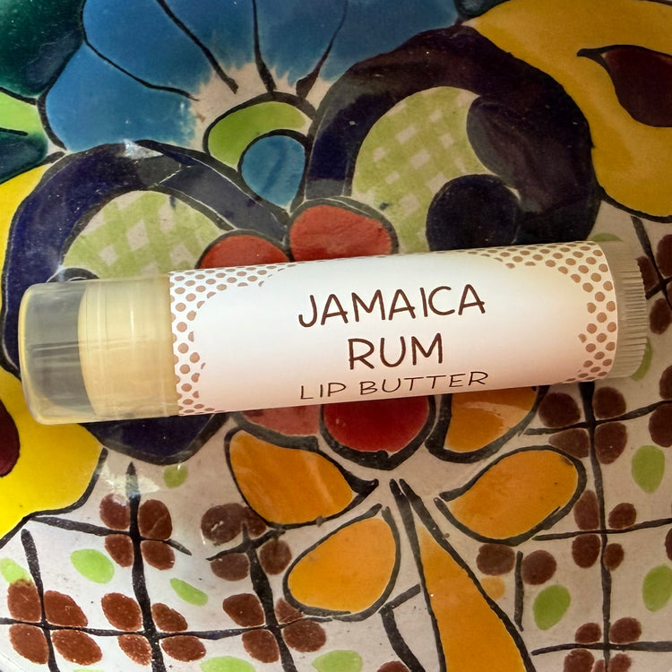 A tube of Jamaica Rum Lip Butter resting on a colorful, hand-painted ceramic tile surface. The lip butter tube has a white label with brown text and polka-dot borders, revealing a pale yellow cream inside.