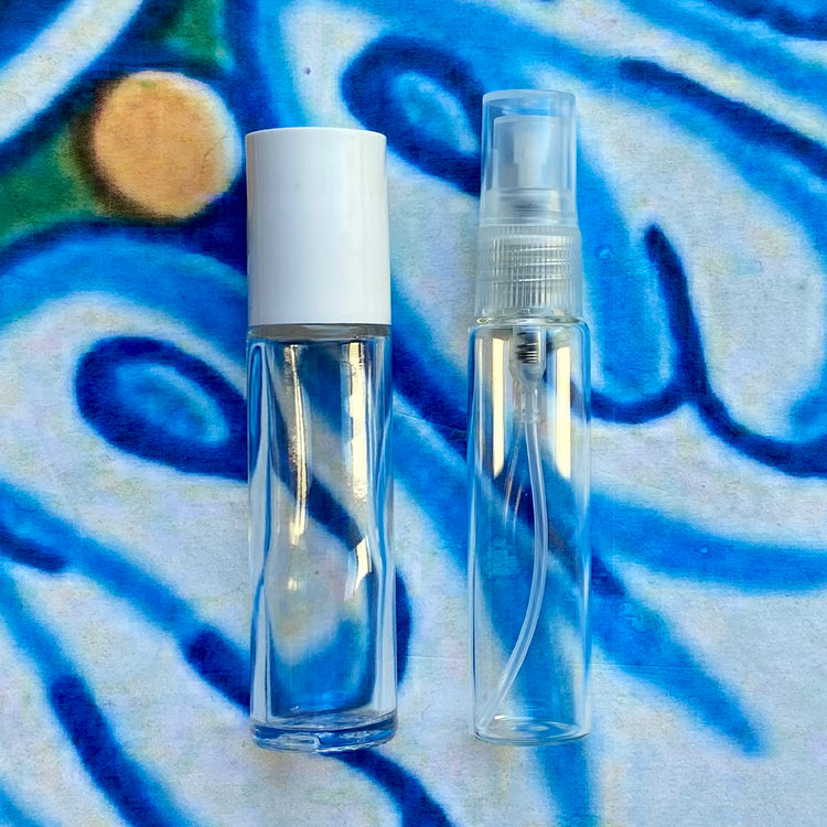 Two perfume containers, one clear with a white cap and one clear with a natural colored top spray, on a colorful abstract background.