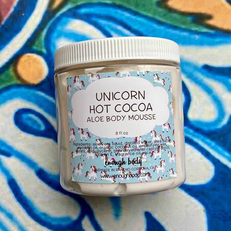 A plastic jar labeled Unicorn Hot Cocoa Aloe Body Mousse on a colorful background.