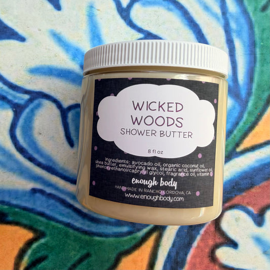 A jar of Enough Body Wicked Woods Shower Butter, 8 fl oz, featuring a white ribbed cap and a black label with a white cloud shape. The product is set against a colorful, patterned background.