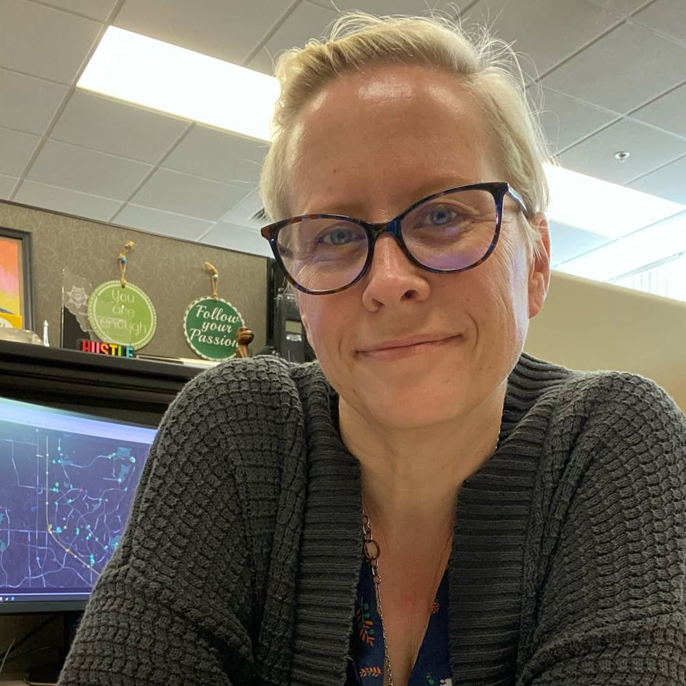 Person wearing glasses and a dark sweater in an office setting with a computer screen in the background.
