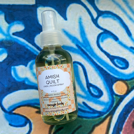 A glass bottle of Amish Quilt Linen and Room Spray with a spray mister top, placed on a surface with a blue and white painted background and floral patterns.