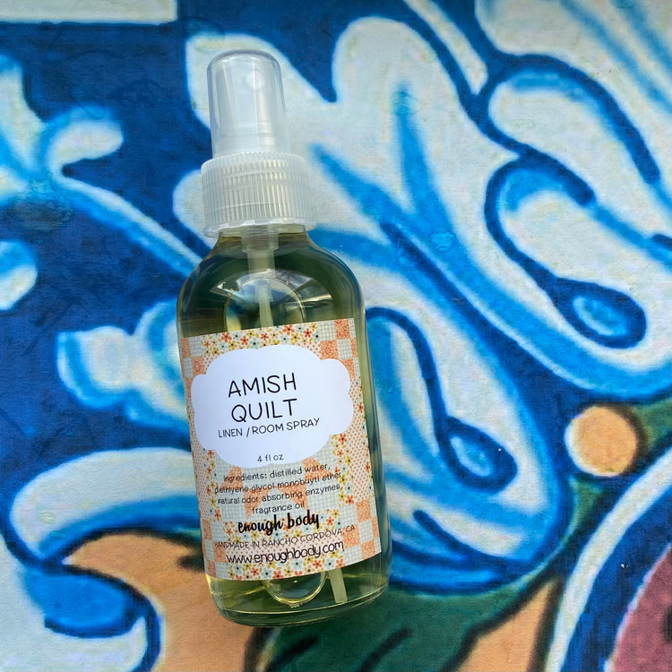 A glass bottle of Amish Quilt Linen and Room Spray with a spray mister top, placed on a surface with a blue and white painted background and floral patterns.