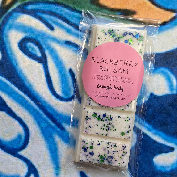 A wax melt snap bar with blackberry balsam label, covered in glitter, placed on a colorful patterned background.
