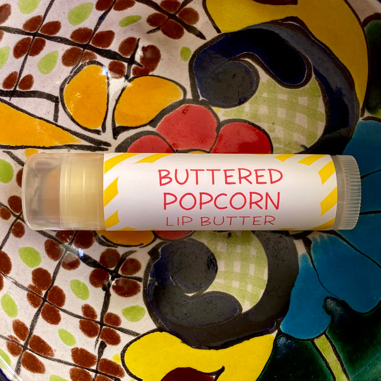 A tube of Buttered Popcorn Lip Butter placed on a colorful patterned surface.