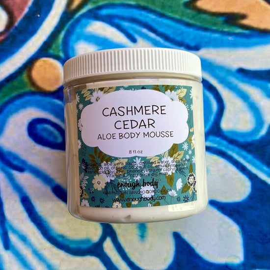 A jar of Cashmere Cedar Aloe Body Mousse with a floral patterned label, placed on a blue patterned background.
