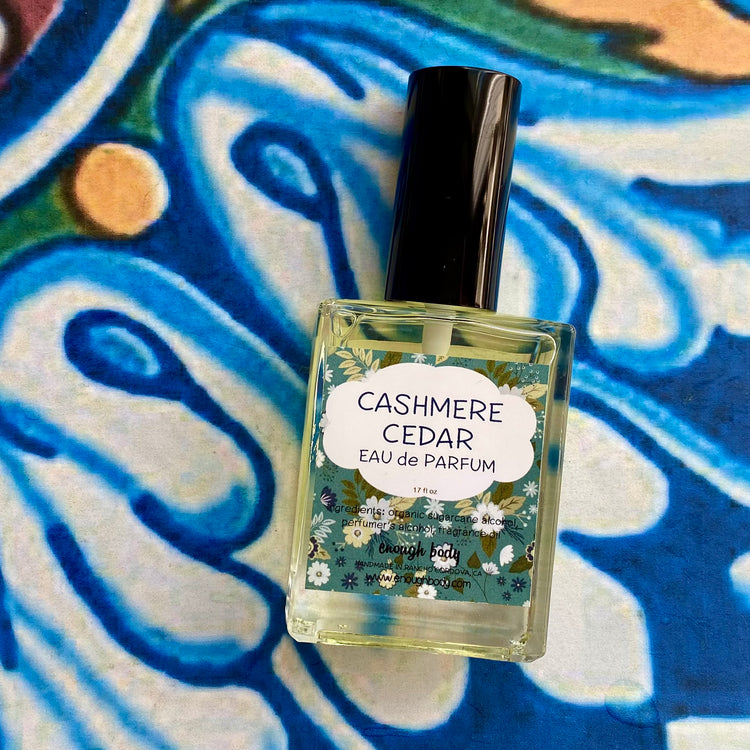 A glass bottle of Cashmere Cedar Eau de Parfum spray with a black spray mister top, labeled with the product name and scent notes, placed on a blue patterned surface.