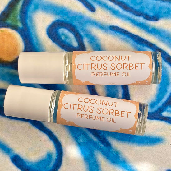 Two glass bottle with a white caps labeled 'coconut citrus sorbet Perfume Oil' set against a blue, floral patterned background.