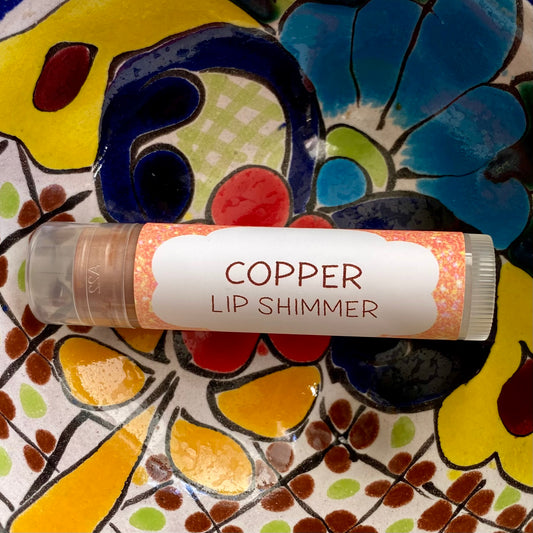 A tube of Copper Lip Shimmer with a colorful background. The tube is clear with a copper-colored lip shimmer visible inside it.