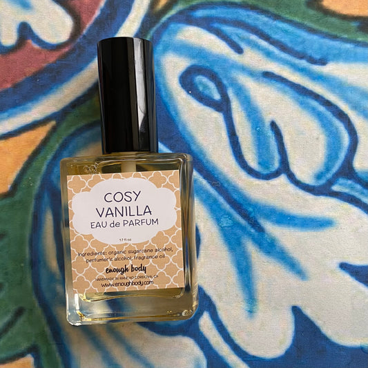 A glass bottle of Cosy Vanilla Eau de Parfum spray with a black spray mister top, placed against a colorful floral background.