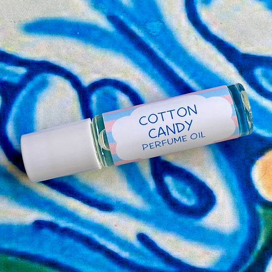A glass bottle with a white cap labeled 'cotton candy perfume oil' set against a blue, floral patterned background.