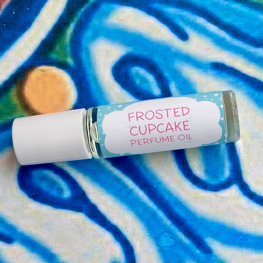 A glass bottle with a white cap labeled 'frosted cupcake perfume oil' set against a blue, floral patterned background.