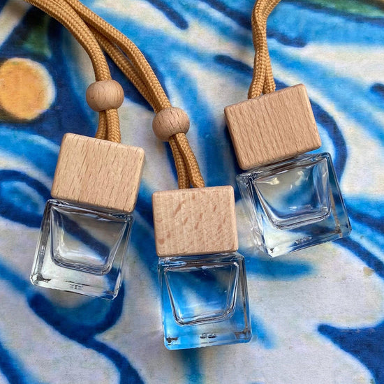 Three square glass car air fresheners with wooden tops on a blue and white patterned background.