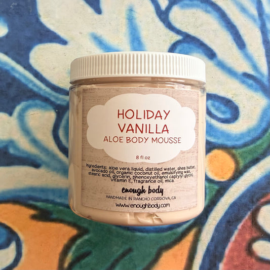 A jar of enough body 'Holiday Vanilla Aloe Body Mousse' on a colorful, patterned ceramic tile background. The product is in a clear plastic jar with a white screw-on lid and a white label with red and black text.