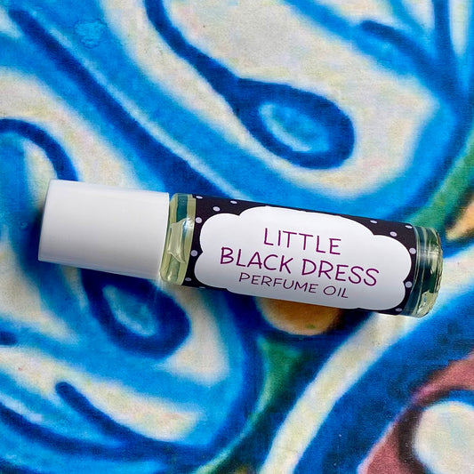 A glass bottle with a white cap labeled 'little black dress perfume oil' set against a blue, floral patterned background.