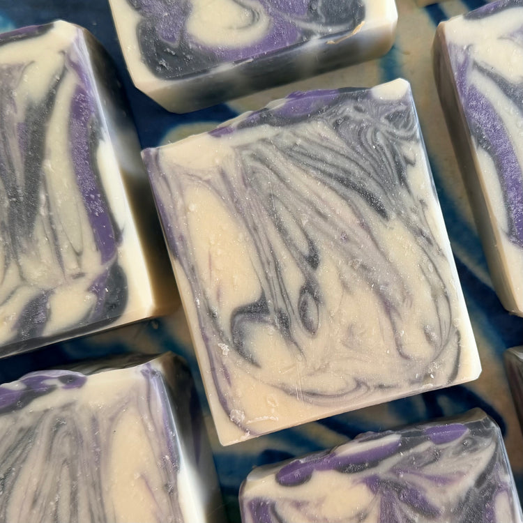 Close-up of several handmade rectangular bars of soap with a marbled swirl pattern in cream, purple, and dark gray, arranged on a blue patterned surface.