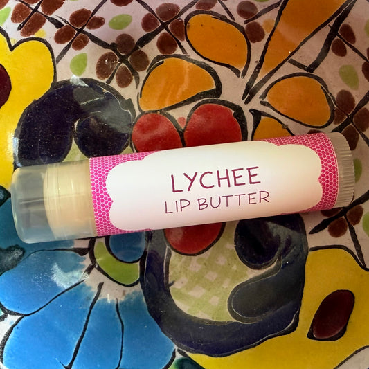 A tube of Lychee Lip Butter resting on a colorful, hand-painted floral surface. The lip balm tube has a white label with pink text and pink patterned borders, and the clear cap reveals a pale yellow balm inside.