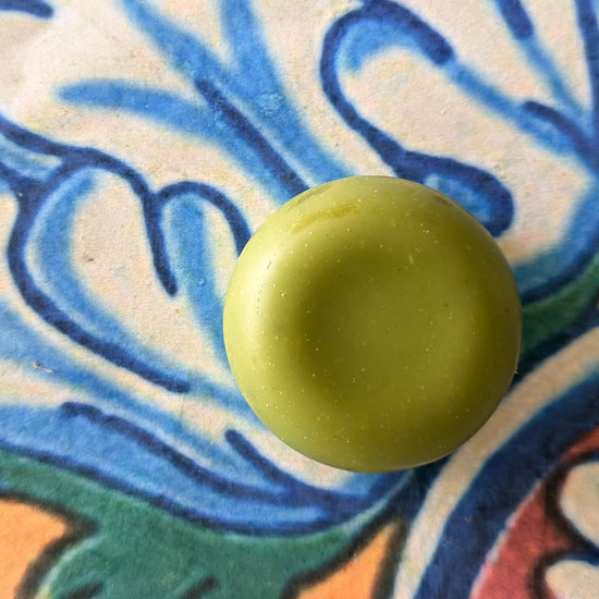 A round, solid bar in a light olive green color, photographed from directly above on a colorful, patterned ceramic tile background.