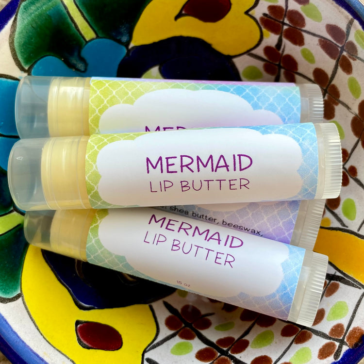 Three tubes of Mermaid Lip Butter with colorful packaging, placed on a ceramic dish with a floral pattern.