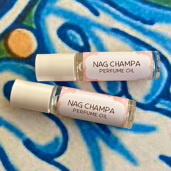 A glass bottle with a white cap labeled 'nag champa Perfume Oil' set against a blue, floral patterned background.