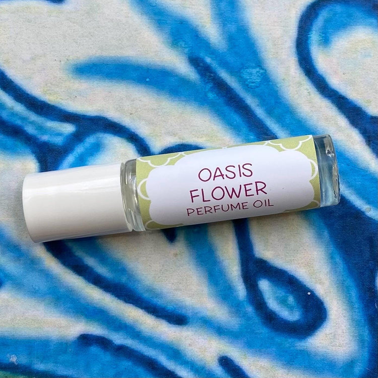 A glass bottle with a white cap labeled 'oasis flower Perfume Oil' set against a blue, floral patterned background.