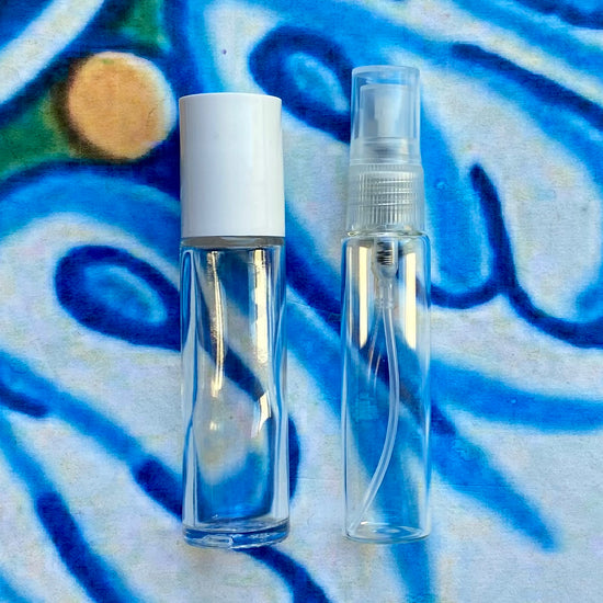 Two perfume containers, one clear with a white cap and one clear with a natural colored top spray, on a colorful abstract background.