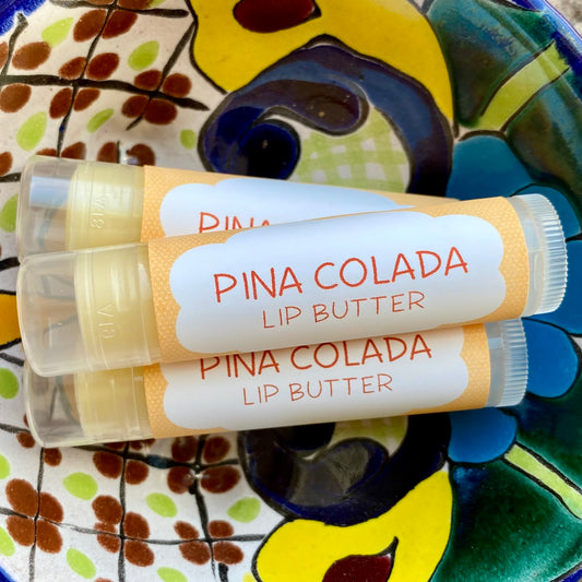 Three tubes of Pina Colada Lip Butter with colorful orange dot packaging, placed on a ceramic dish with a floral pattern.