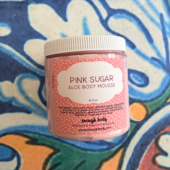 A jar of enough body 'Pink Sugar Aloe Body Mousse' in a pink container with a white lid, set against a colorful blue, white, and orange patterned tile background.