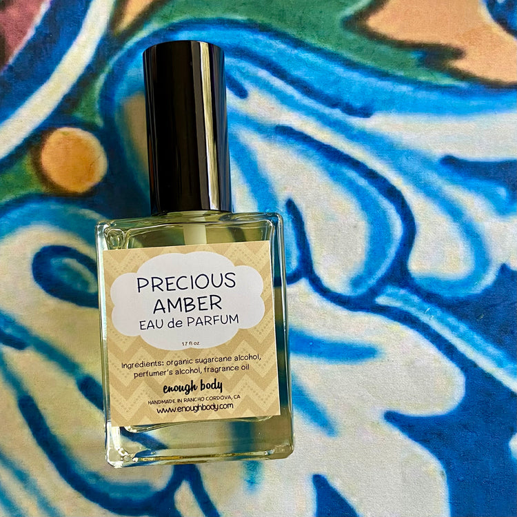 A glass bottle of Precious Amber Eau de Parfum spray with a label and a black spray mister top, placed on a patterned fabric.