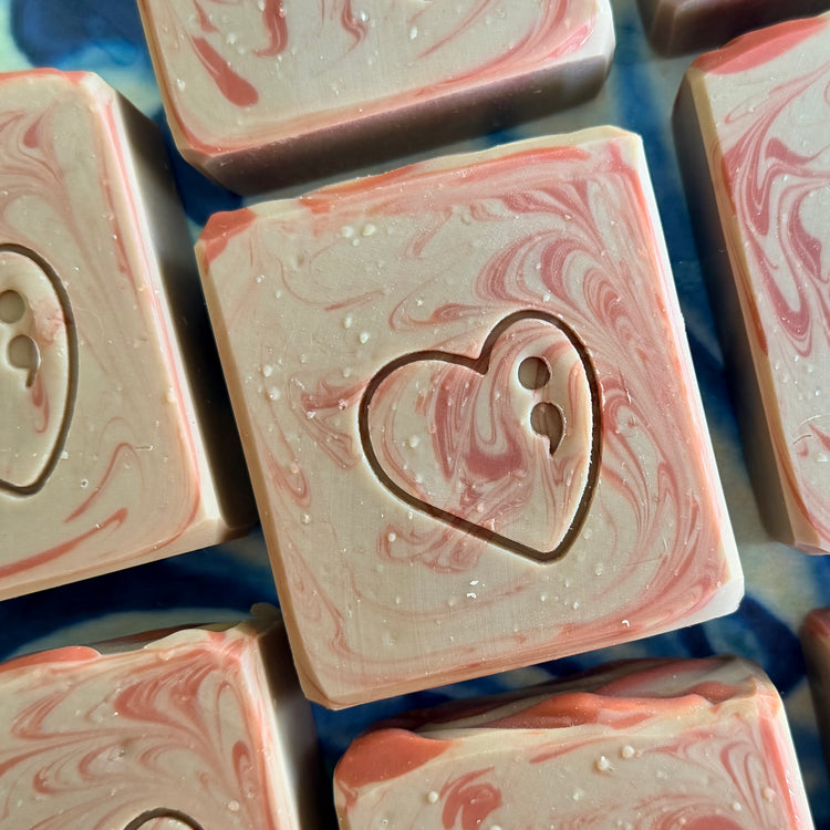 Close-up of several square handmade soap bars with pink and cream marbled swirls, one centered bar featuring an embossed heart with two dots, resting on a blue patterned surface.