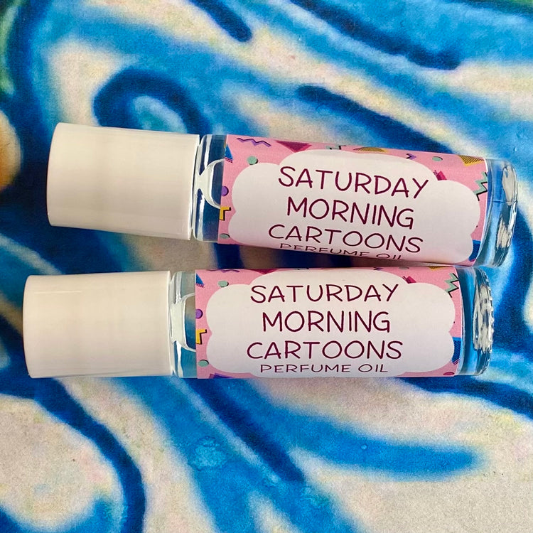 Two bottles of 'Saturday Morning Cartoons' perfume oil with label that smells like cartoons', placed on a patterned fabric. The bottles are clear with a white cap and label that has the product name and a cartoon graphic.