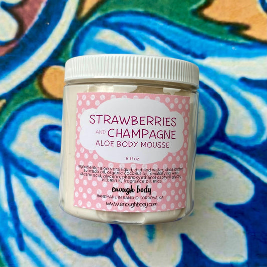A jar of Strawberries and Champagne Aloe Body Mousse with a label showing the product name and a polka dot pattern. The jar is on a background with a colorful, floral pattern.