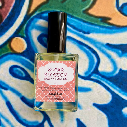 A glass bottle of Sugar Blossom Eau de Parfum spray with a black spray mister top, labeled with a floral design, placed on a colorful patterned background.