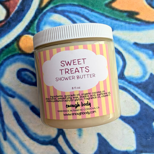 A jar of enough body Sweet Treats Shower Butter sits on a colorful, patterned surface. The product is in a clear plastic jar showing the beige shower butter inside, with a white screw-on lid and a pink and yellow striped label.