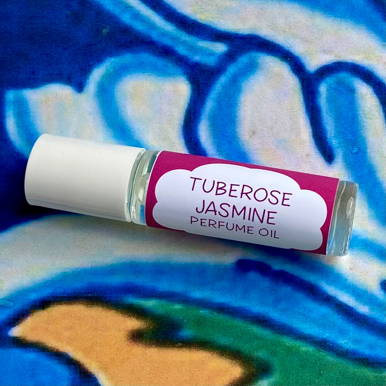A glass bottle with a white cap labeled 'tuberose jasmine Perfume Oil' set against a blue, floral patterned background.