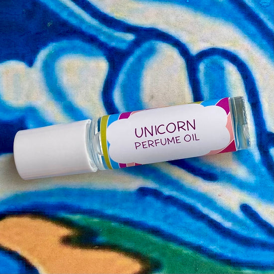 A glass bottle with a white cap labeled 'unicorn perfume oil' set against a blue, floral patterned background.