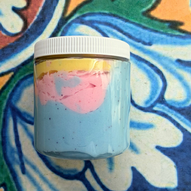 A clear plastic jar filled with layered cream or butter, showing a light blue base, a pink middle layer, and a yellow top layer, sealed with a white ribbed screw cap, set against a colorful patterned background.