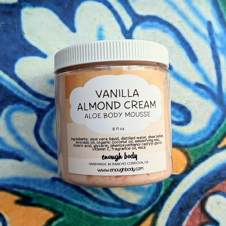 A jar of enough body Vanilla Almond Cream Aloe Body Mousse, featuring a white lid and a beige label, set against a vibrant blue and white patterned tile background.