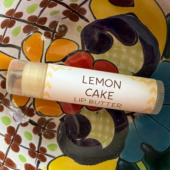 A tube of Lemon Cake Lip Butter resting on a colorful, patterned ceramic plate. The lip butter packaging features a white label with brown text and yellow chevron accents.