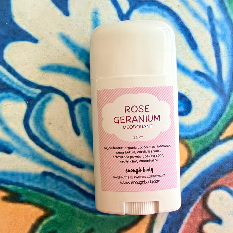 A white plastic deodorant stick labeled 'Rose Geranium' by enough body, placed on a colorful patterned fabric background.