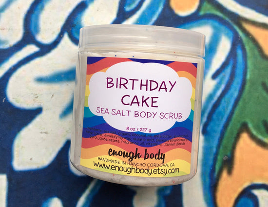 A plastic jar of 'Birthday Cake Sea Salt Body Scrub' with colorful jojoba beads visible through the clear jar, set against a patterned background.