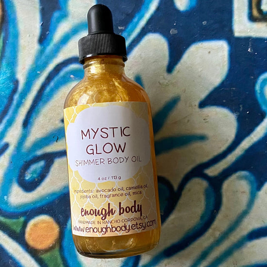 A glass bottle with a black dropper filled with yellow body oil, labeled 'Mystic Glow Shimmer Body Oil' on a colorful patterned background.